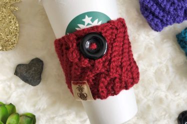 A crochet coffee cozy made with the Kelsi Cozy crochet pattern in red with large black button