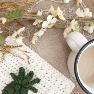 An off-white crochet square that is part of a blanket CAL with a dark green crochet pineapple applique sitting atop a neutral background accented by white flowers and coffee mug.