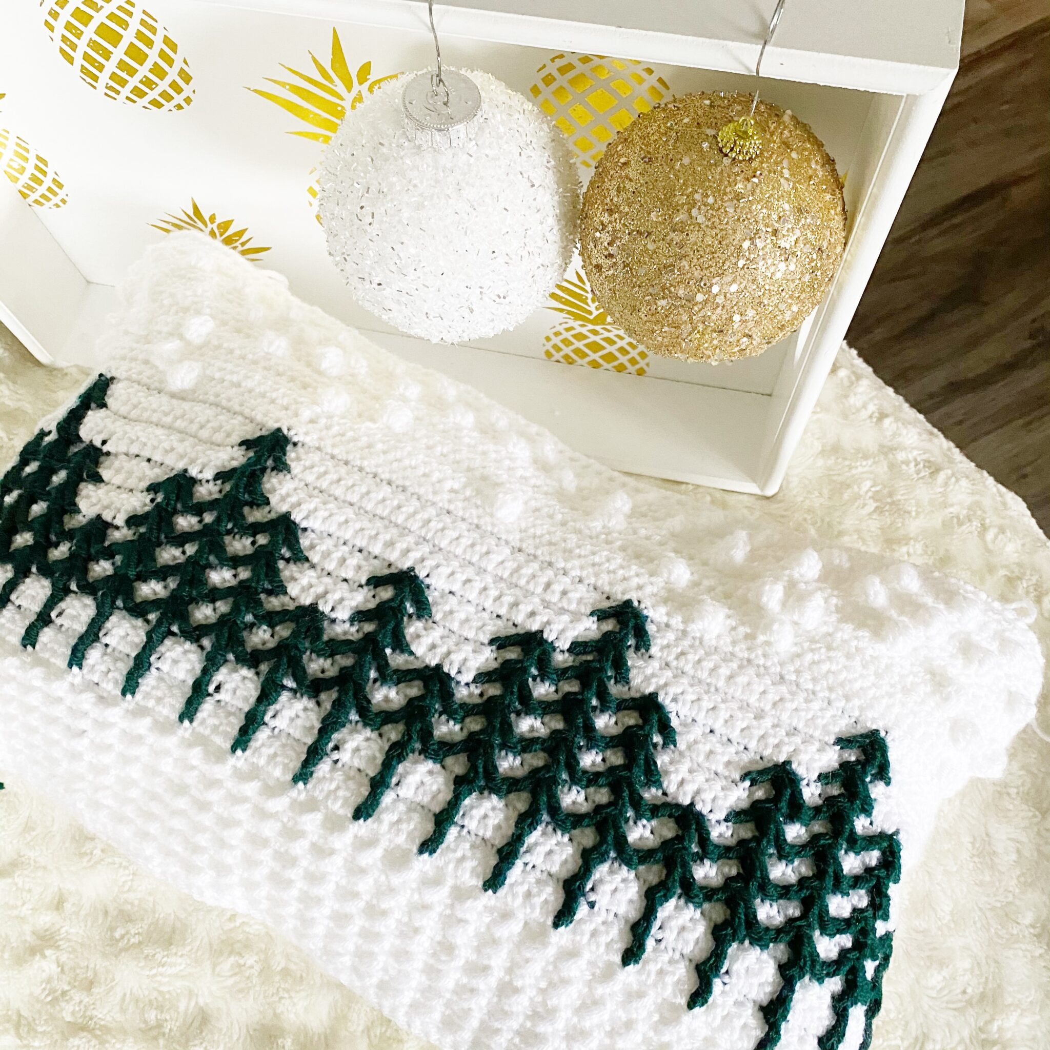 crochet holiday pillow featuring white background with green pine trees and white bobbles that represent falling snow
